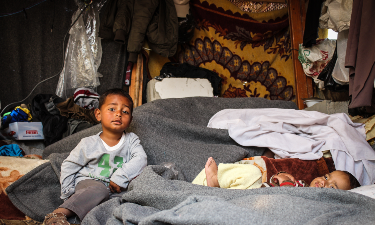 Two small children resting inside a makeshift shelter — the kind of home the orphanage appeal aims to replace